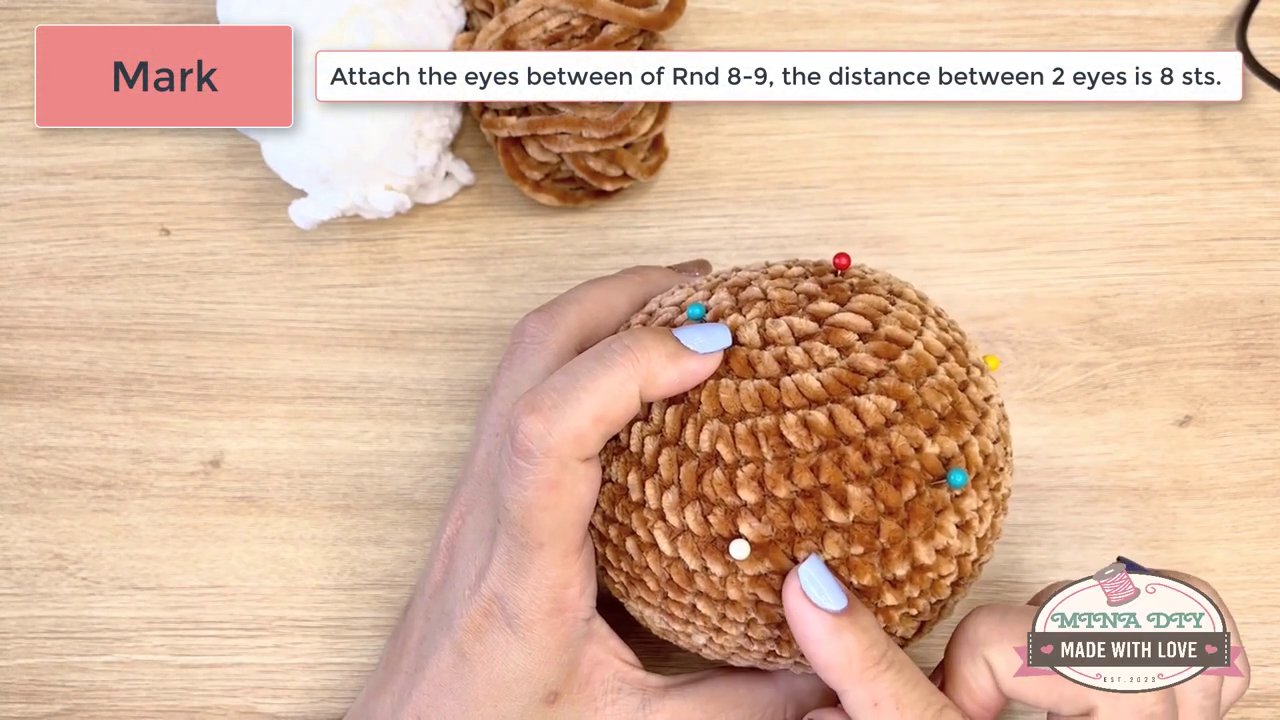 Bringing Your Amigurumi to Life: A Step-by-Step Guide to Embroidering Eyes and Noses 1 A pair of hands with blue and white painted nails gently shapes the face of a brown crocheted animal on a wooden crafting table.